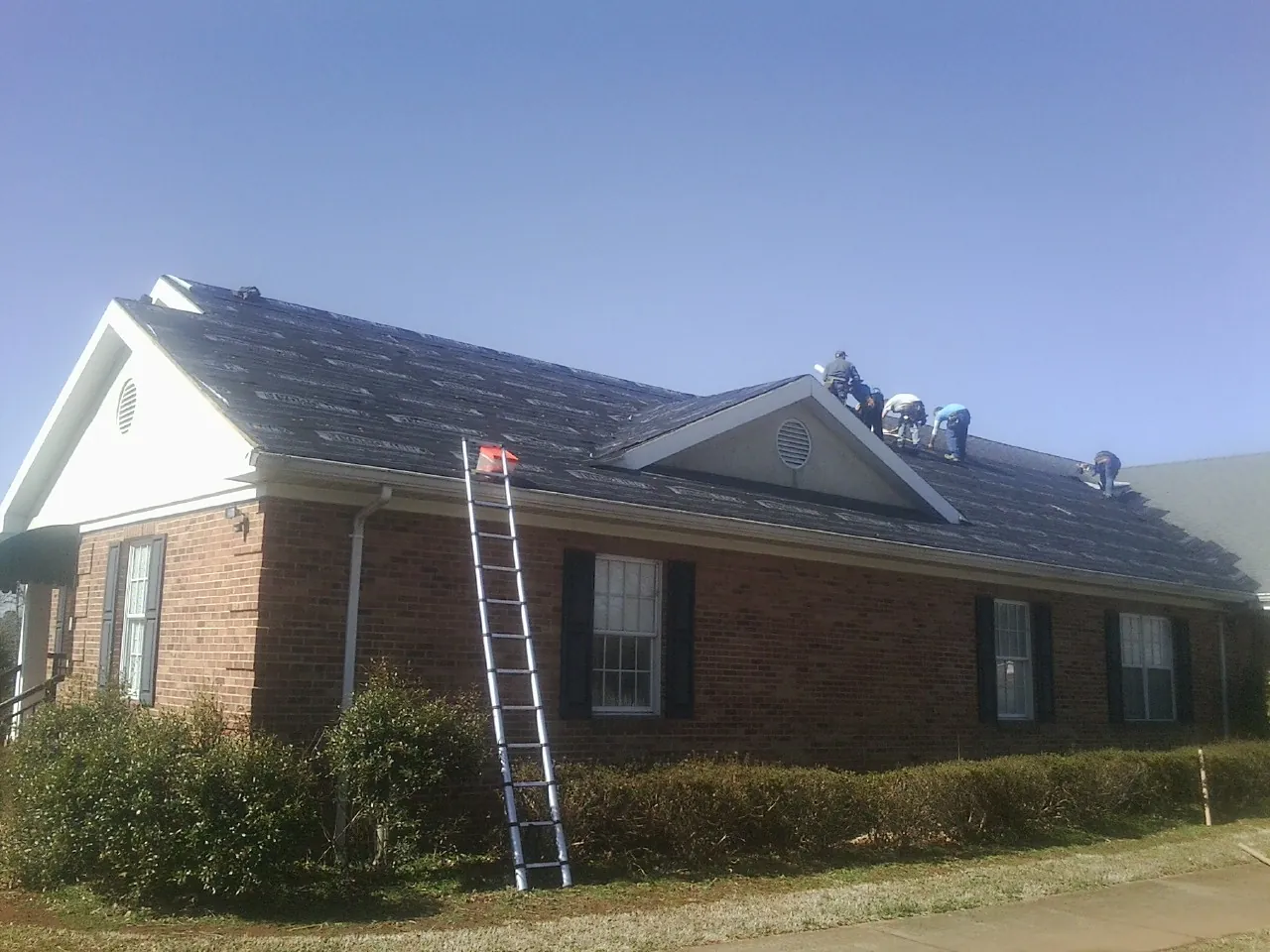 Crew tearing shingles off a brick ranch roof with a ladder set — Emergency Roof Repair work by our crew in Sugar Land