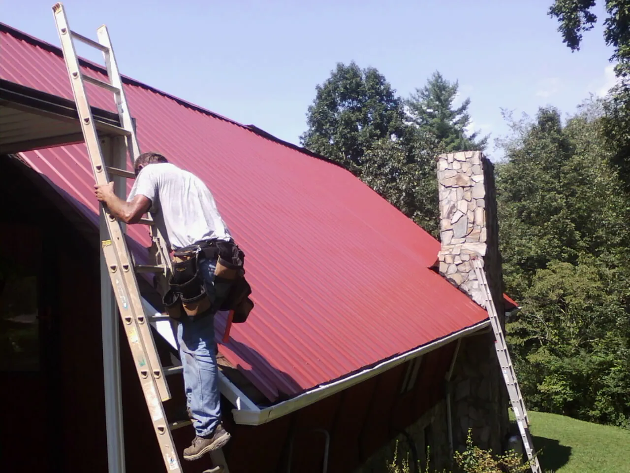 Roofer on a ladder beside a red metal roof and stone chimney — Soffit & Fascia Repair by Roofing Brothers in Sugar Land